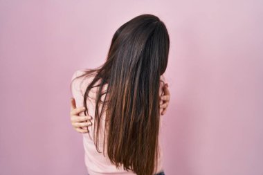Genç esmer kadın pembe arka planda dikilip kendine sarılıyor mutlu ve pozitif bir şekilde. Sevgi ve özgüven. 