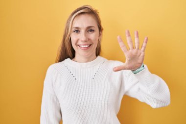 Beyaz süveter giyen, sarı arka planda beş numaralı parmaklarıyla gülümseyen, kendinden emin ve mutlu beyaz bir kadın.. 