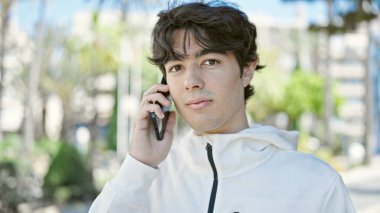 Young hispanic man talking on smartphone with serious expression at park