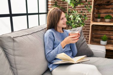 Genç sarışın kadın evde kahve içip kitap okuyor.