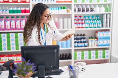 Young beautiful hispanic woman pharmacist reading notebook using computer at pharmacy