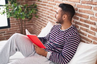 Young hispanic man reading book sitting on bed at bedroom