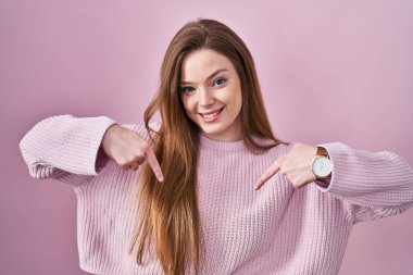 Pembe arka planda duran, kendine güvenen, kendini gururlu ve mutlu gösteren genç, beyaz bir kadın.. 