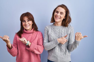 Anne ve kızı mavi arka planda dikiliyorlar. Elleri ve başparmakları havada, kendinden emin gülümsüyorlar. 