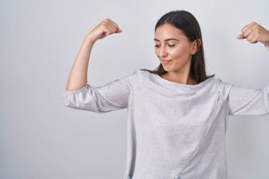 Young hispanic woman standing over white background showing arms muscles smiling proud. fitness concept. 