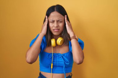 Sarı arka planda duran Latin kökenli genç bir kadın, elleri başının üstünde, stresten baş ağrısı çekiyor. migren ağrısı. 
