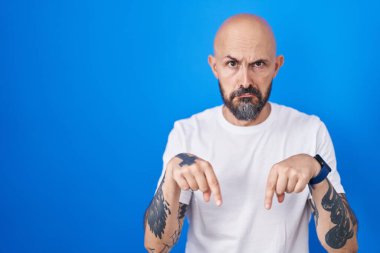 Mavi arka planda dikilen İspanyol dövmeli adam üzgün ve üzgün görünüyor, parmaklarıyla yönü gösteriyor, mutsuz ve depresif.. 