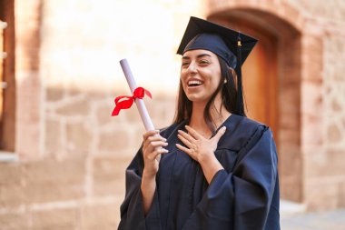 Young hispanic woman wearing graduated uniform holding diploma at university