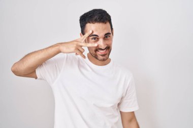 Beyaz arka planda duran yakışıklı İspanyol adam barış sembolü yapıyor. Yüzünde parmakları, gülümseyerek zaferi gösteriyor. 