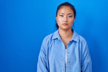 Mavi arka planda duran Asyalı genç kadın yüzünde ciddi bir ifadeyle rahatlamış. Basit ve doğal bir şekilde kameraya bakıyorsun.. 