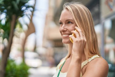 Genç sarışın kadın gülümsüyor. Kendine güveni tam. Akıllı telefondan konuşuyor.