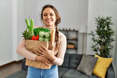 Genç, güzel, İspanyol bir kadın gülümsüyor. Elinde alışveriş torbası var.
