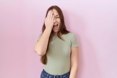 Güzel esmer kadın pembe arka planda duruyor. Bir gözünü eliyle kapatıyor. Yüzünde kendine güvenen bir gülümseme ve sürpriz bir duygu var.. 