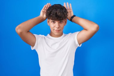 Latin kökenli genç, mavi arka planda durup, avuçlarında alaycı ve şüpheci bakışlarla tavşan kulağı hareketi yapıyor. Paskalya tavşanı konsepti. 