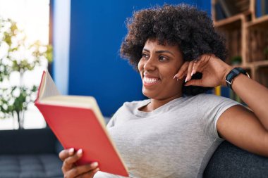 Evdeki kanepede oturan Afro-Amerikalı kadın kitap okuyor.