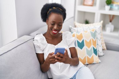 Evdeki koltukta oturan Afro-Amerikan kadın akıllı telefon kullanıyor