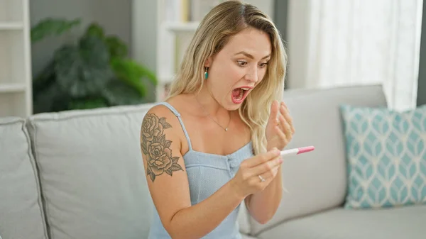 Young blonde woman holding pregnancy test with surprise expression at home
