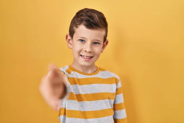 黄色い背景の上に立っている若い慎重な子供は 挨拶と歓迎としてハンドシェイクを提供しています 成功したビジネス — ストック写真