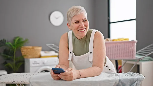 Middle age grey-haired woman using smartphone leaning on ironing board smiling at laundry room