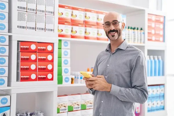 Young hispanic man customer smiling confident using smartphone at pharmacy