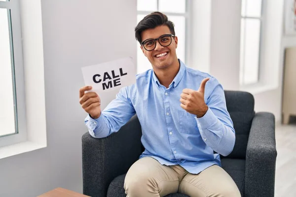 Hispanic man working at therapy office holding call me banner smiling happy and positive, thumb up doing excellent and approval sign