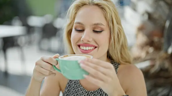 Young blonde woman smelling cup of coffee sitting on table at coffee shop terrace