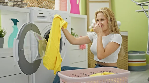 洗濯室で汚れたTシャツを嗅ぐ若い金髪の女性 — ストック写真