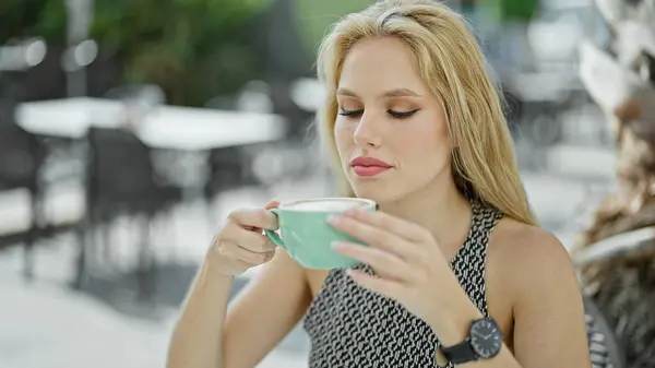 Young blonde woman holding cup of coffee sitting on table at coffee shop terrace