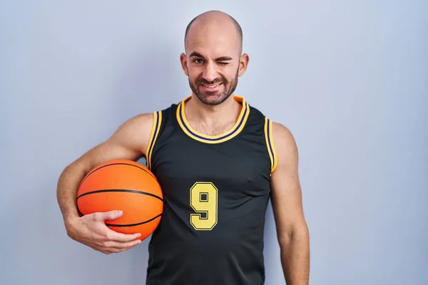 年轻的秃头男子 留着胡子 身穿篮球服 手握球 眨着眼睛 带着性感的表情 笑容满面地看着摄像机 — 图库照片