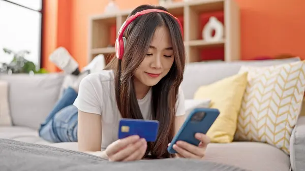 Young chinese woman shopping with smartphone and credit card listening to music at home