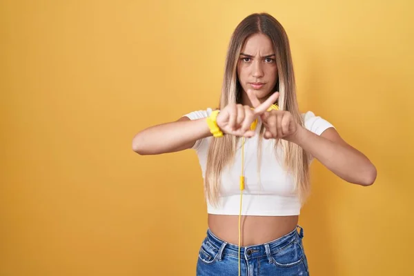 年轻的金发女人站在黄色的背景上 头戴耳机 戴着拒收的表情 手指交叉 做着消极的手势 — 图库照片