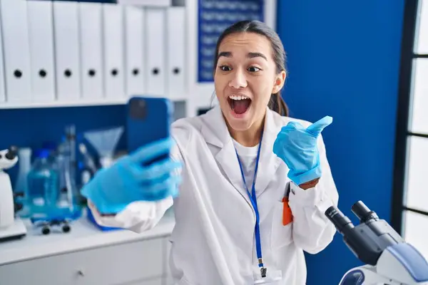 Young south asian woman working at scientist laboratory with smartphone pointing thumb up to the side smiling happy with open mouth