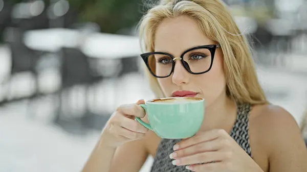 Young blonde woman drinking cup of coffee sitting on table thinking at coffee shop terrace