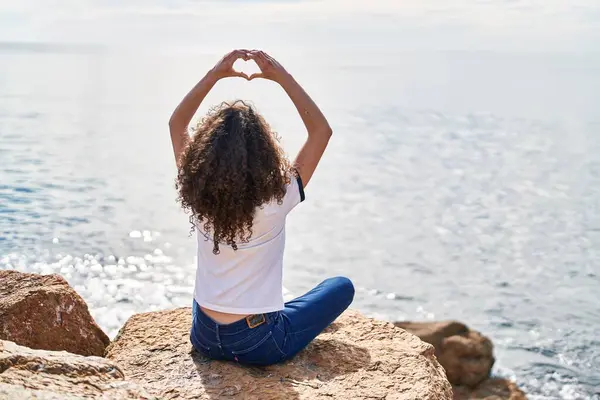 Young hispanic woman sitting on back view doing heart gesture with hands at seaside
