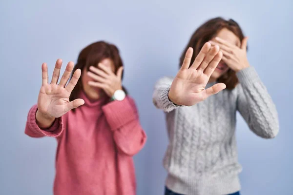 Mother and daughter standing over blue background covering eyes with hands and doing stop gesture with sad and fear expression. embarrassed and negative concept.