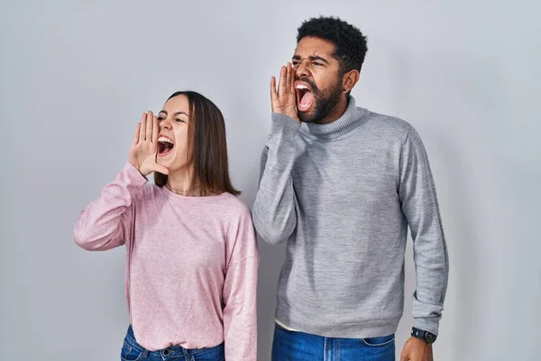 Young hispanic couple standing together shouting and screaming loud to side with hand on mouth. communication concept.