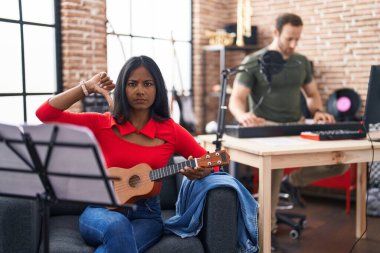 Genç Hintli bir kadın müzik stüdyosunda ukulele çalıyor. Kızgın bir yüzü var. Eksi işareti baş parmakları aşağıda, reddedilme konsepti ise... 