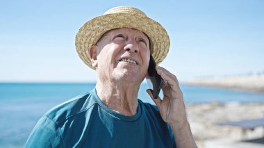 Kıdemli kır saçlı turist. Yazlık şapka takıyor. Deniz kenarında akıllı telefondan konuşuyor.