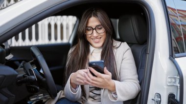 Young beautiful hispanic woman using smartphone sitting on car at street