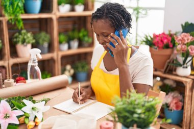 Orta yaşlı Afro-Amerikan kadın çiçekçi çiçekçi çiçekçide defterde akıllı telefon yazısıyla konuşuyor.