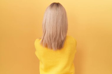 Sarı süveter giyen genç beyaz kadın kollarını kavuşturup arkasını kolluyor. 