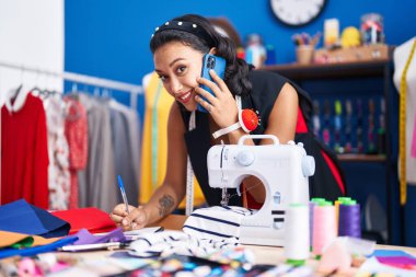 Young beautiful hispanic woman tailor talking on smartphone writing on notebook at clothing factory