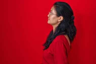 Asyalı genç bir kadın kırmızı arka planda duruyor, yan tarafa bakıyor, rahat poz veriyor doğal bir yüz ve kendine güvenen bir gülümsemeyle.. 