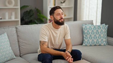 Kanepede oturan Latin asıllı genç adam. Ciddi bir suratı var.