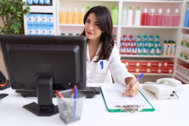 Middle age hispanic woman pharmacist writing on document using computer at pharmacy