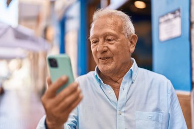 Yaşlı, gri saçlı, kendine güvenen, caddede akıllı telefon kullanan bir adam.