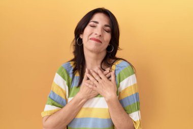 Middle age hispanic woman standing over yellow background smiling with hands on chest with closed eyes and grateful gesture on face. health concept. 