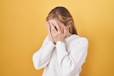 Sarı arka plan üzerine beyaz kazak giyen beyaz bir kadın. Ağlarken suratını ellerle kapatan üzgün bir ifade. depresyon kavramı. 