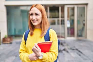 Üniversitede elinde kitaplarla gülümseyen beyaz bir bayan öğrenci.