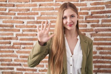 Kafkasyalı genç bir kadın duvarın arkasında duruyor. Arka planda beş numarayı gösteriyor ve işaret ediyor. Kendinden emin ve mutlu bir şekilde gülümsüyor.. 
